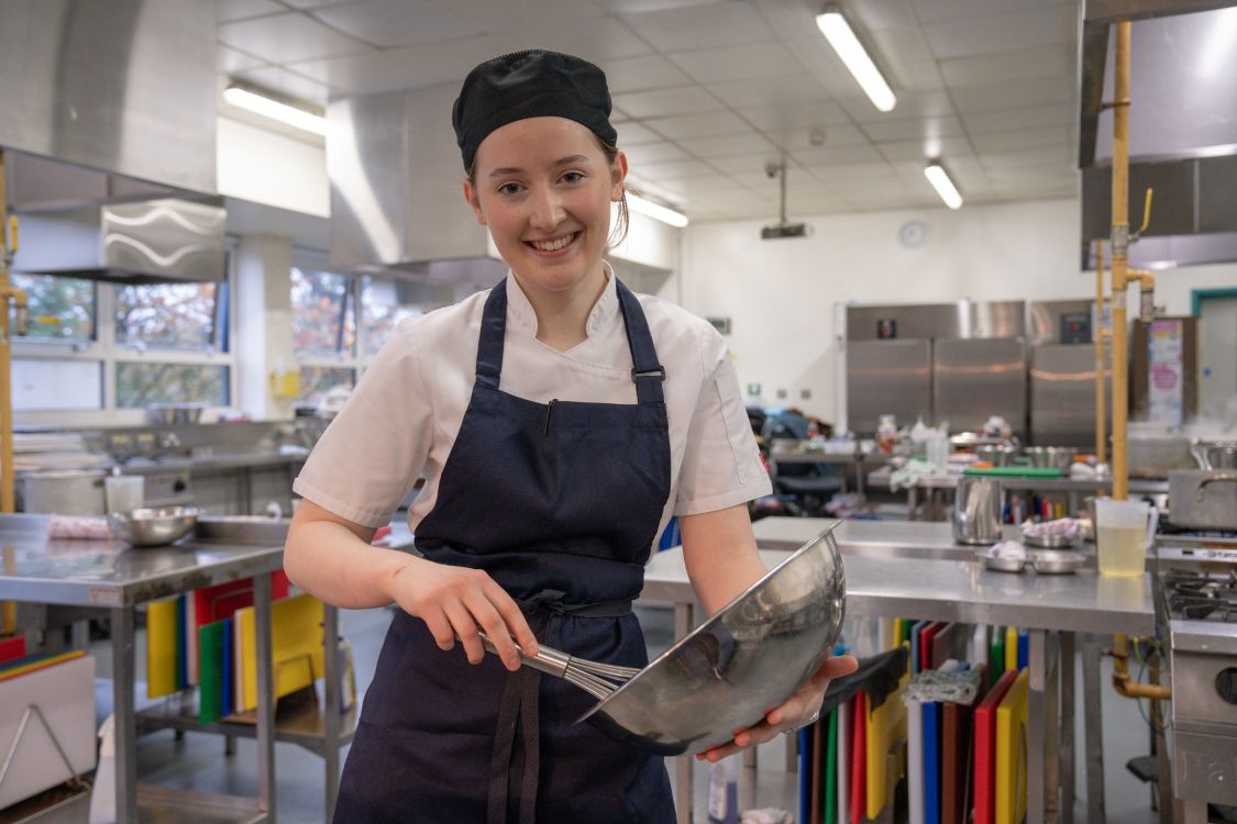 A young woman in an apron and hat with a mixing bowl and whisk