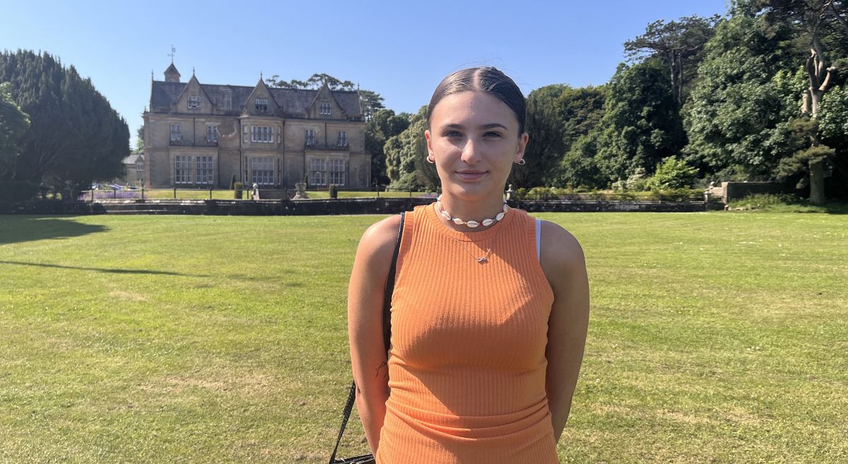 Ali McKay-Geary, young woman with dark hair wearing orange top pictured in bright sunshine in front of historic building with large lawn